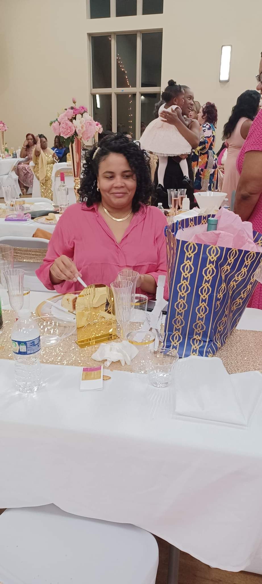 Woman in pink shirt at decorated table, smiling. Other people and decorations in background.