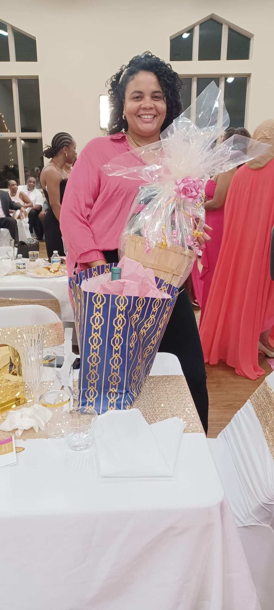 Woman smiles, holding gift basket; wearing pink shirt, patterned pants. Event setting.