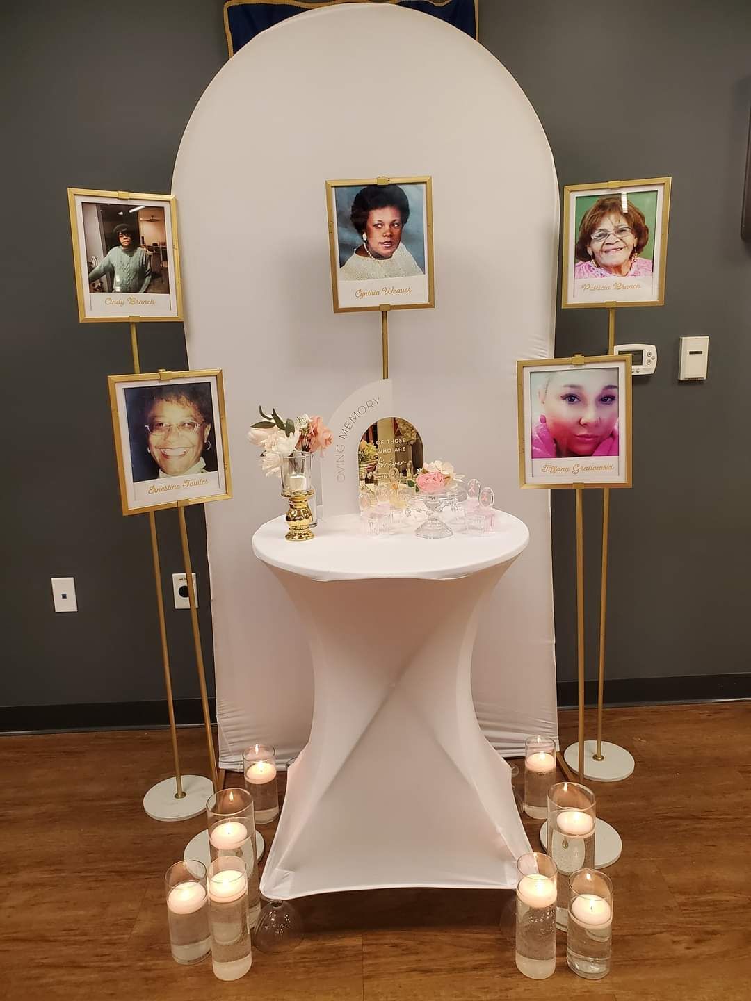 Memorial with photos, candles, and flowers on a table. Gold frames with portraits around a white arch and table.