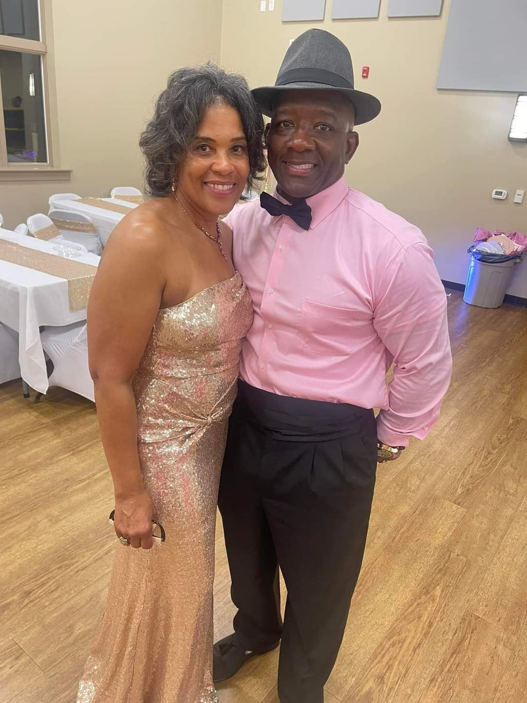 Couple at event, woman in gold gown, man in pink shirt and hat smiling indoors.