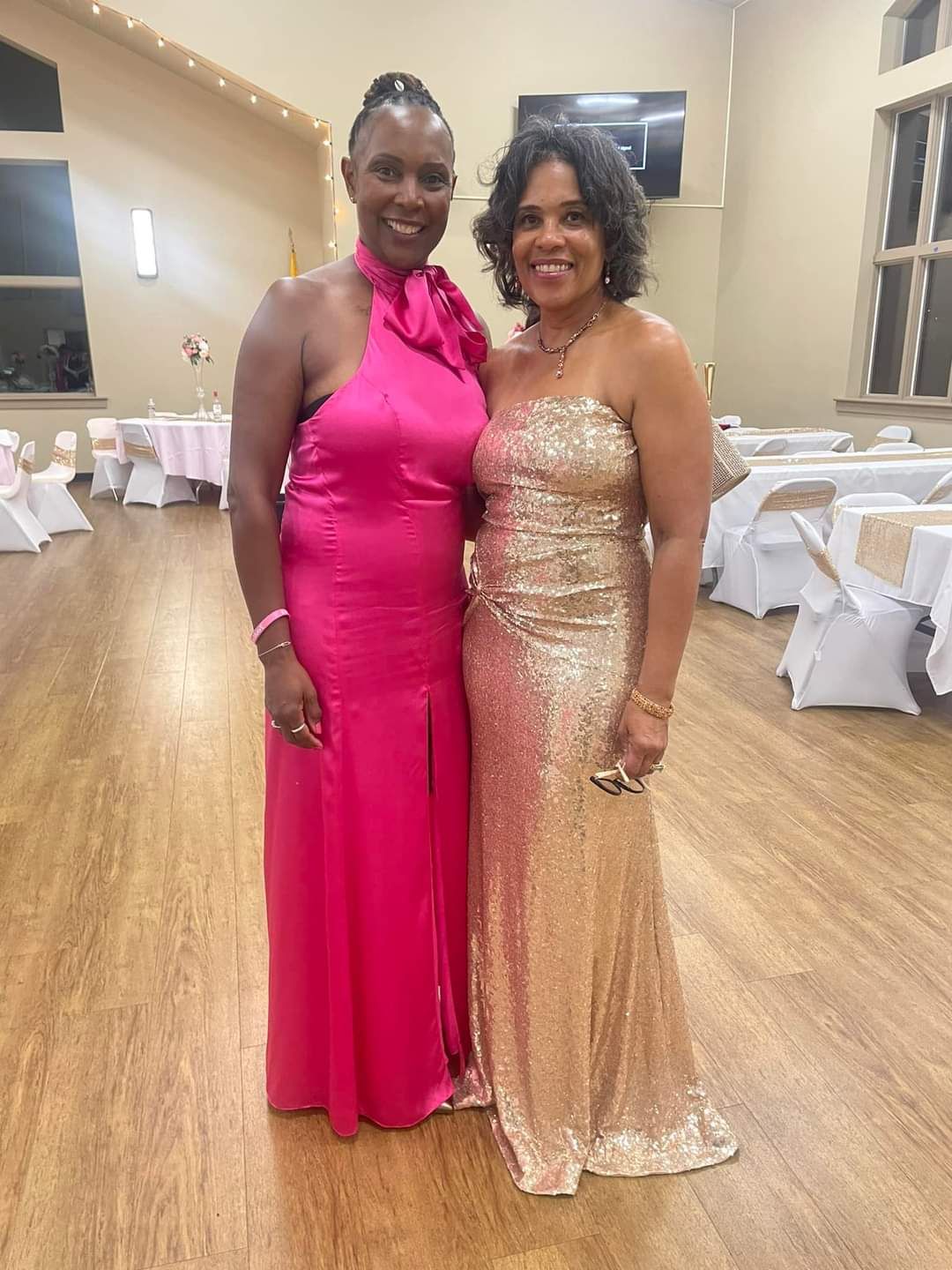 Two women in formal gowns at an event; one in pink, the other in gold. They smile at the camera.