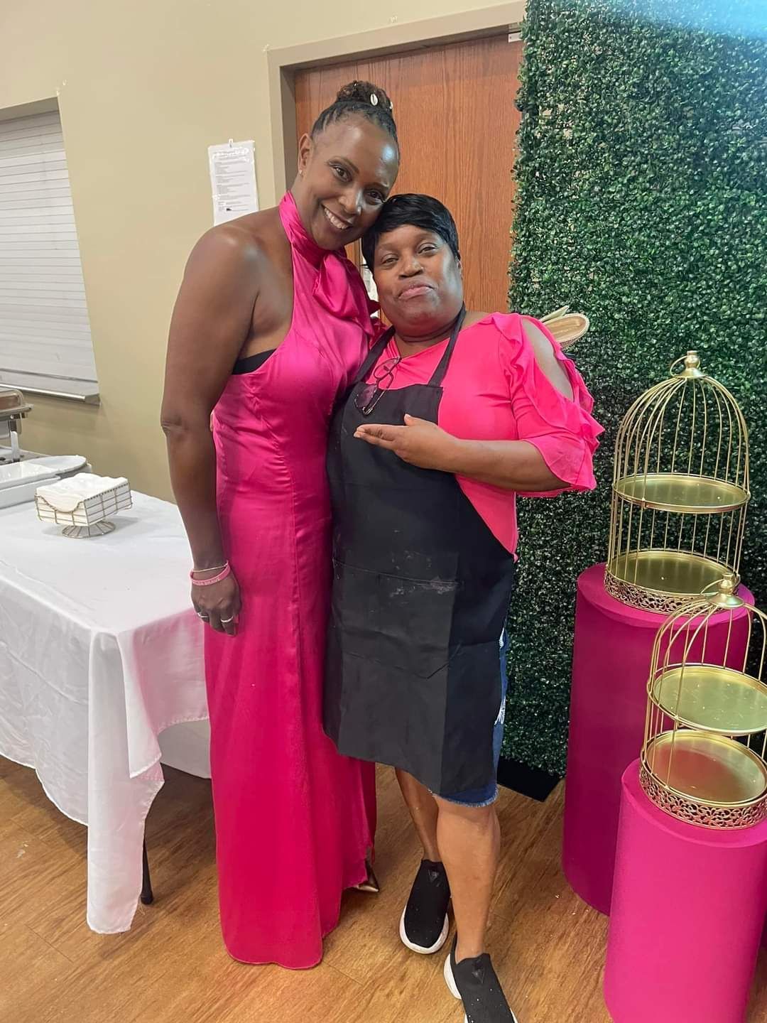 Two women pose together: one in a pink dress, the other in an apron.