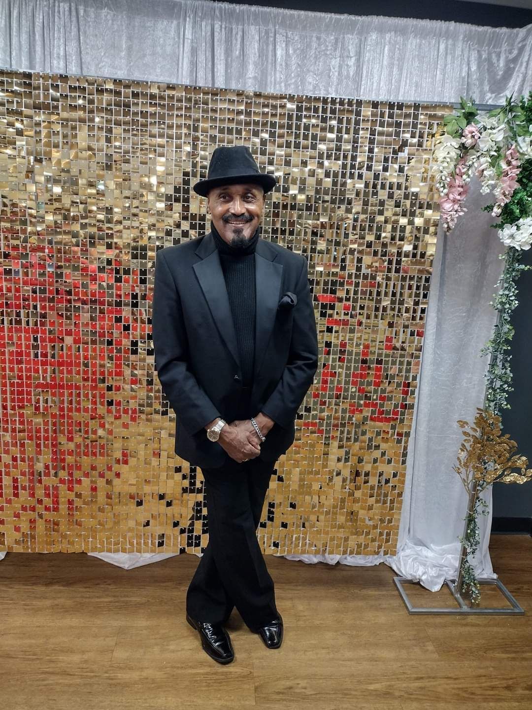 Man in black suit and hat, smiling, posing in front of a gold sequin backdrop.