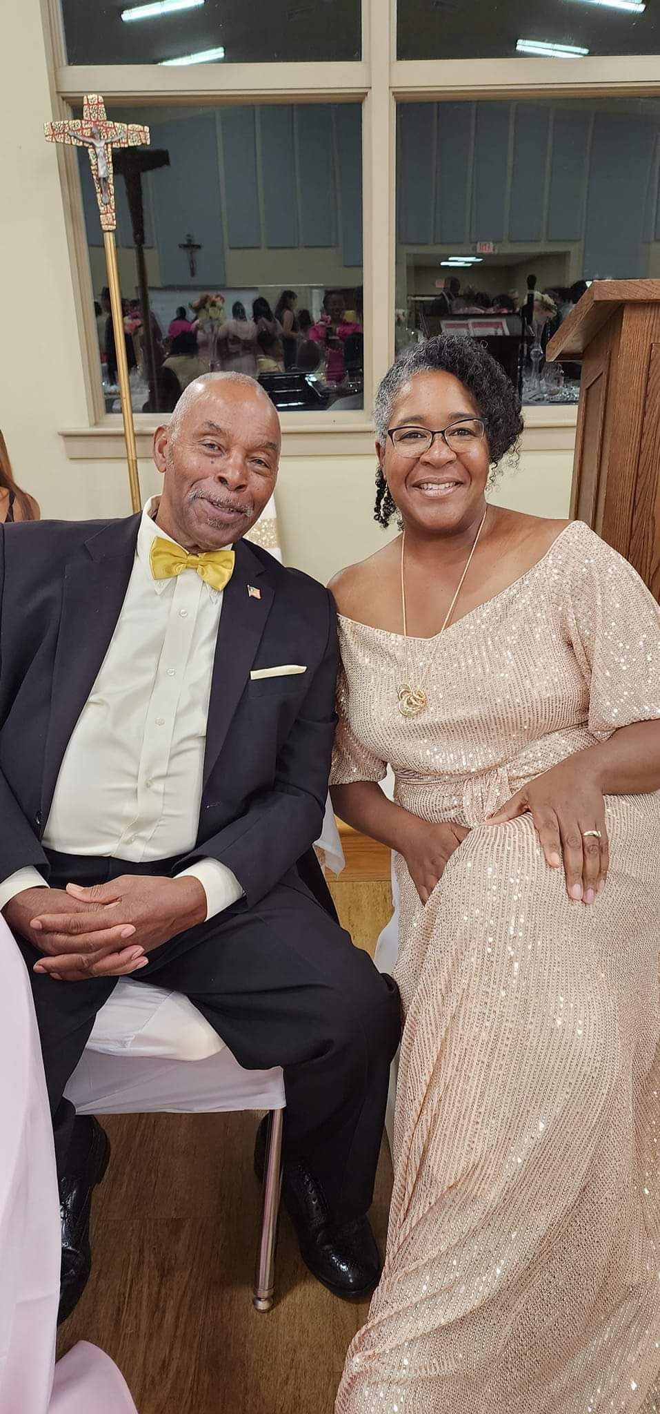 An elegantly dressed couple smiles. Man in suit and yellow bow tie; woman in sequined gown. Indoors, church setting.