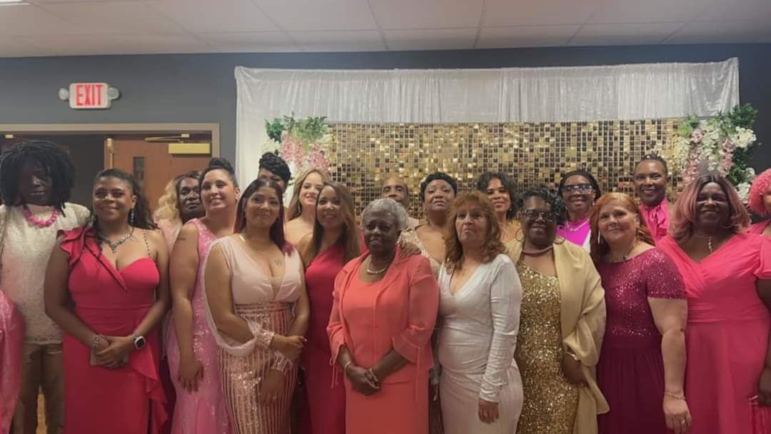 Group of women in pink and gold dresses posing in front of a gold and white backdrop.