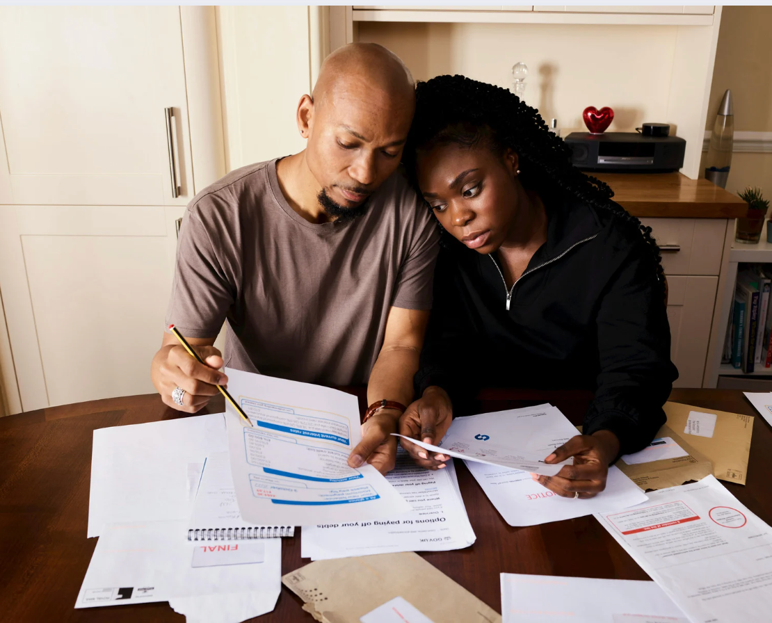 Couple reviewing bills at a table, appearing stressed. Papers, pen, and printer visible.
