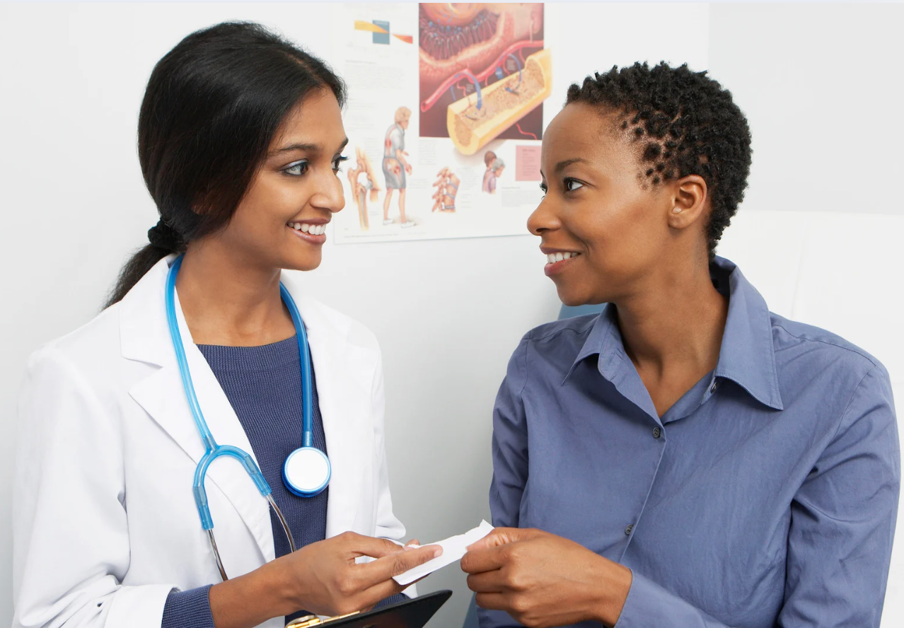 Doctor in white coat giving a prescription to a patient. Both women smile in a medical office.