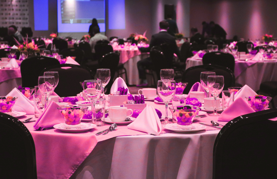 Formal event with round tables set for dinner; pink and white decor, people in suits.