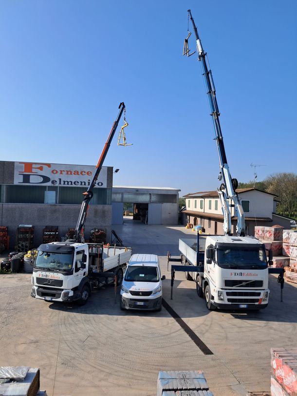 Due autogru, un furgone bianco e un edificio sotto un cielo azzurro. I camion sollevano oggetti.