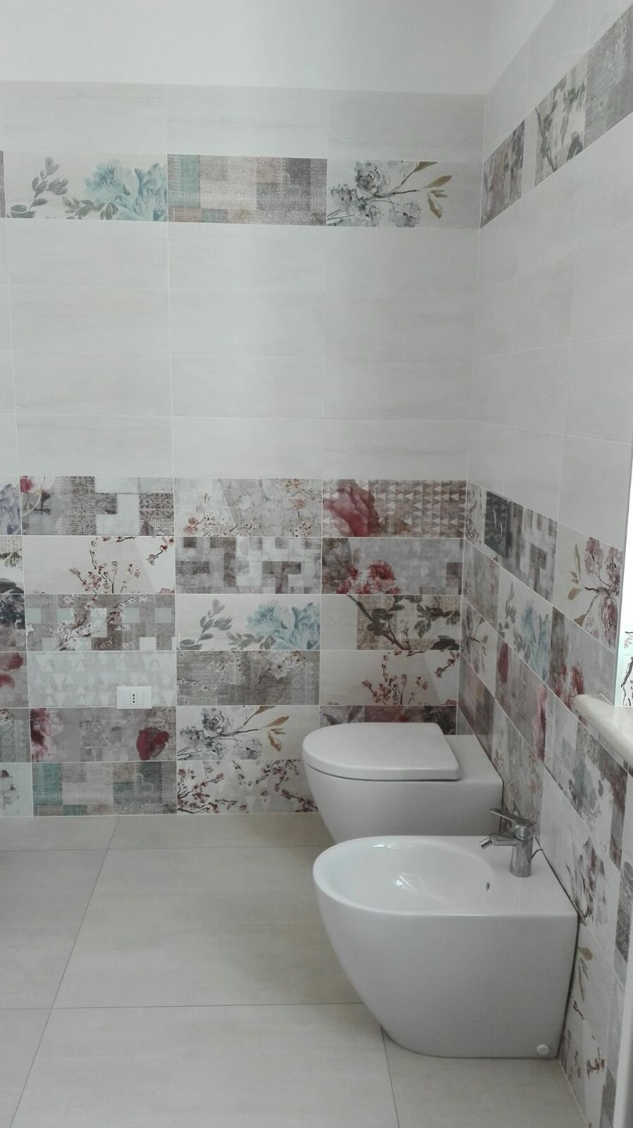Bagno con sanitari bianchi, piastrelle decorative con motivi floreali e semplici piastrelle bianche alle pareti.
