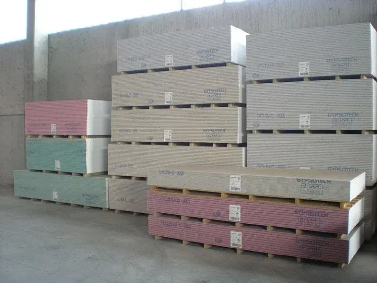 Pile di lastre di cartongesso su pallet di legno in un magazzino; alcune lastre sono rosa o verdi.