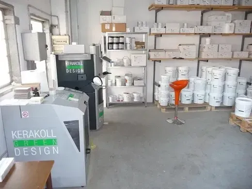 Interno di un laboratorio Kerakoll Design con macchinari, scaffali con prodotti e uno sgabello arancione.