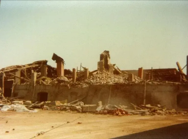 Detriti di un edificio crollato; tetto e pareti distrutti, restano solo i camini in mattoni. Fuori, giornata luminosa.