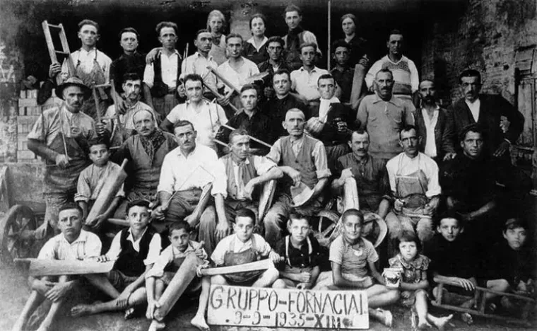 Un gruppo di uomini e ragazzi posa, tenendo in mano attrezzi e legna, con l'insegna