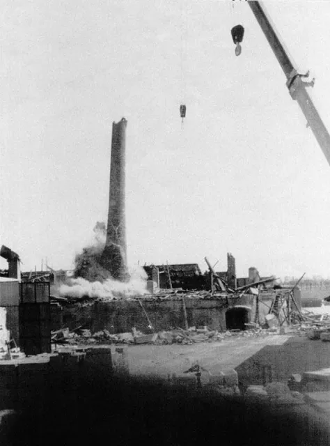 Demolizione di un edificio in mattoni e della sua ciminiera, con gru, detriti e polvere.