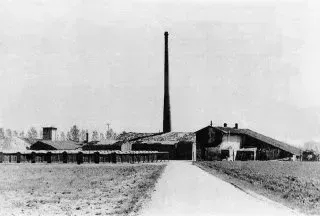 Foto in bianco e nero di un lungo edificio con un'alta ciminiera; sembra un sito industriale o una fabbrica.