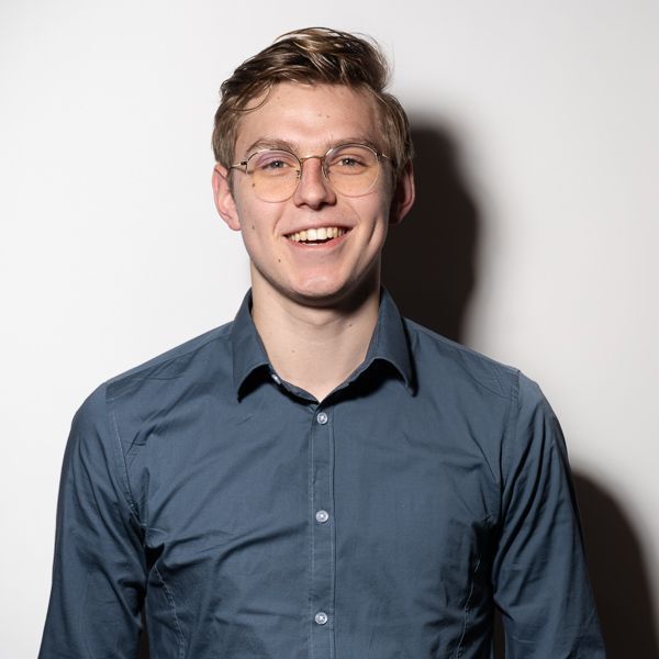 Een jonge man met een bril en een blauw shirt lacht naar de camera