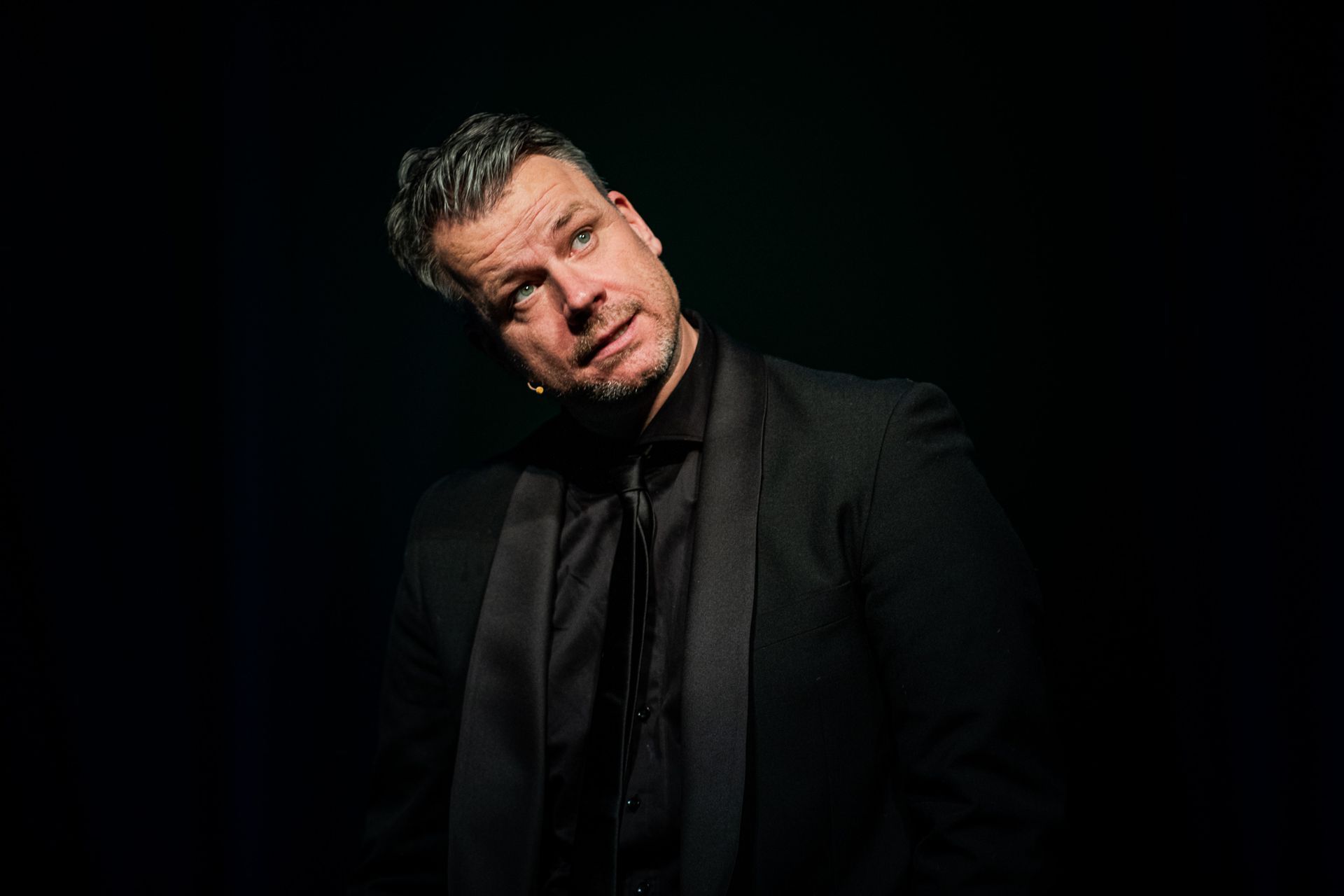 Een man in een zwart pak en stropdas trekt een raar gezicht.