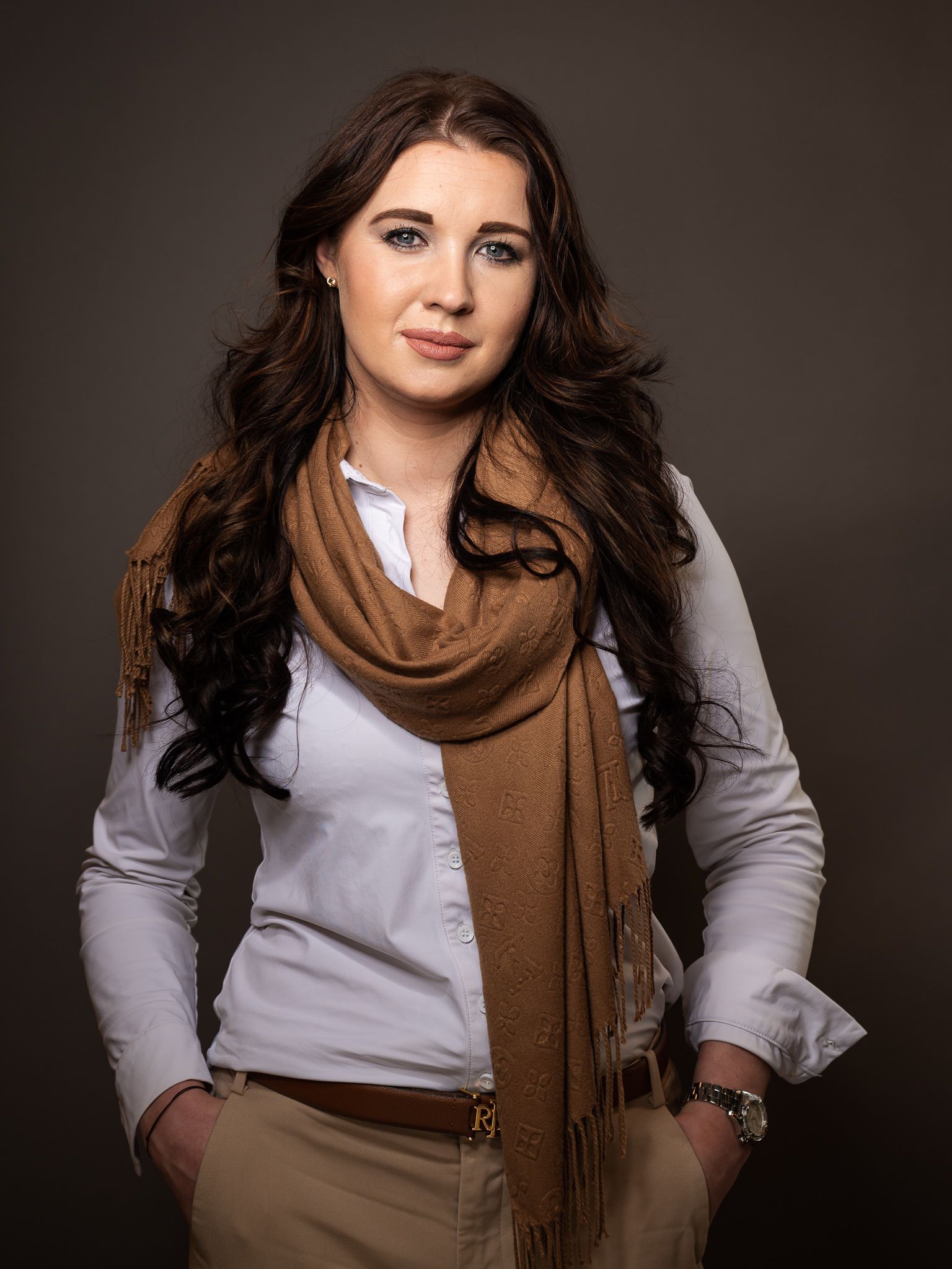 Een vrouw draagt ​​een wit shirt en een bruine sjaal en staat met haar handen in haar zakken.