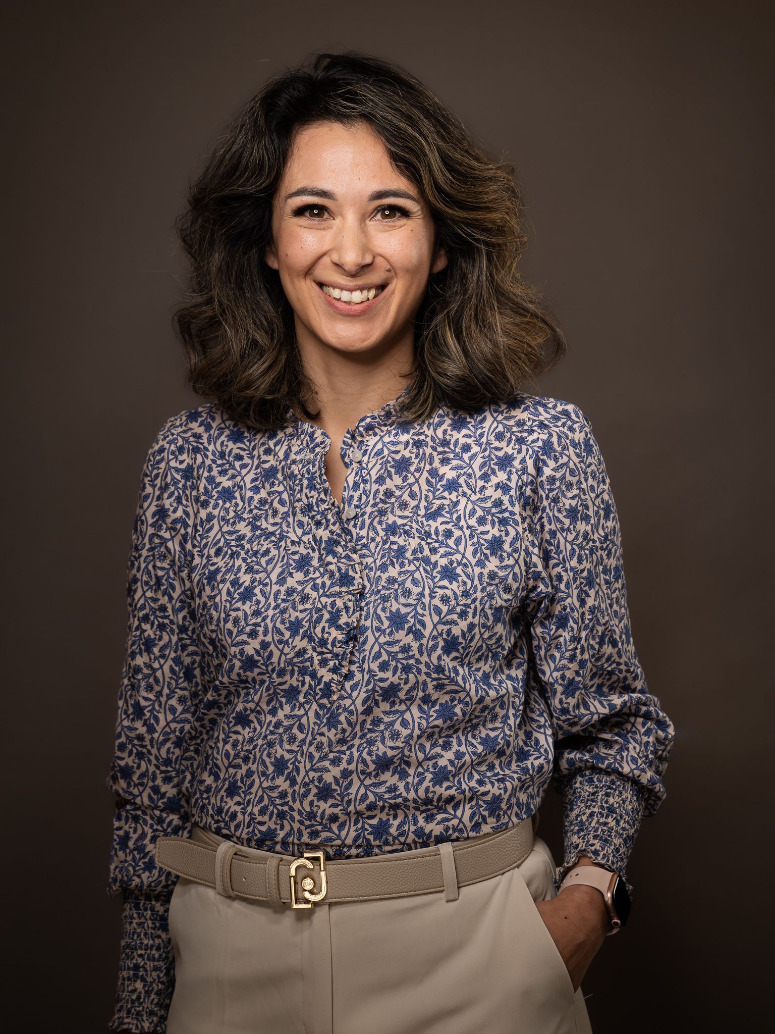 Een vrouw in een blauw shirt en een kaki broek lacht naar de camera.