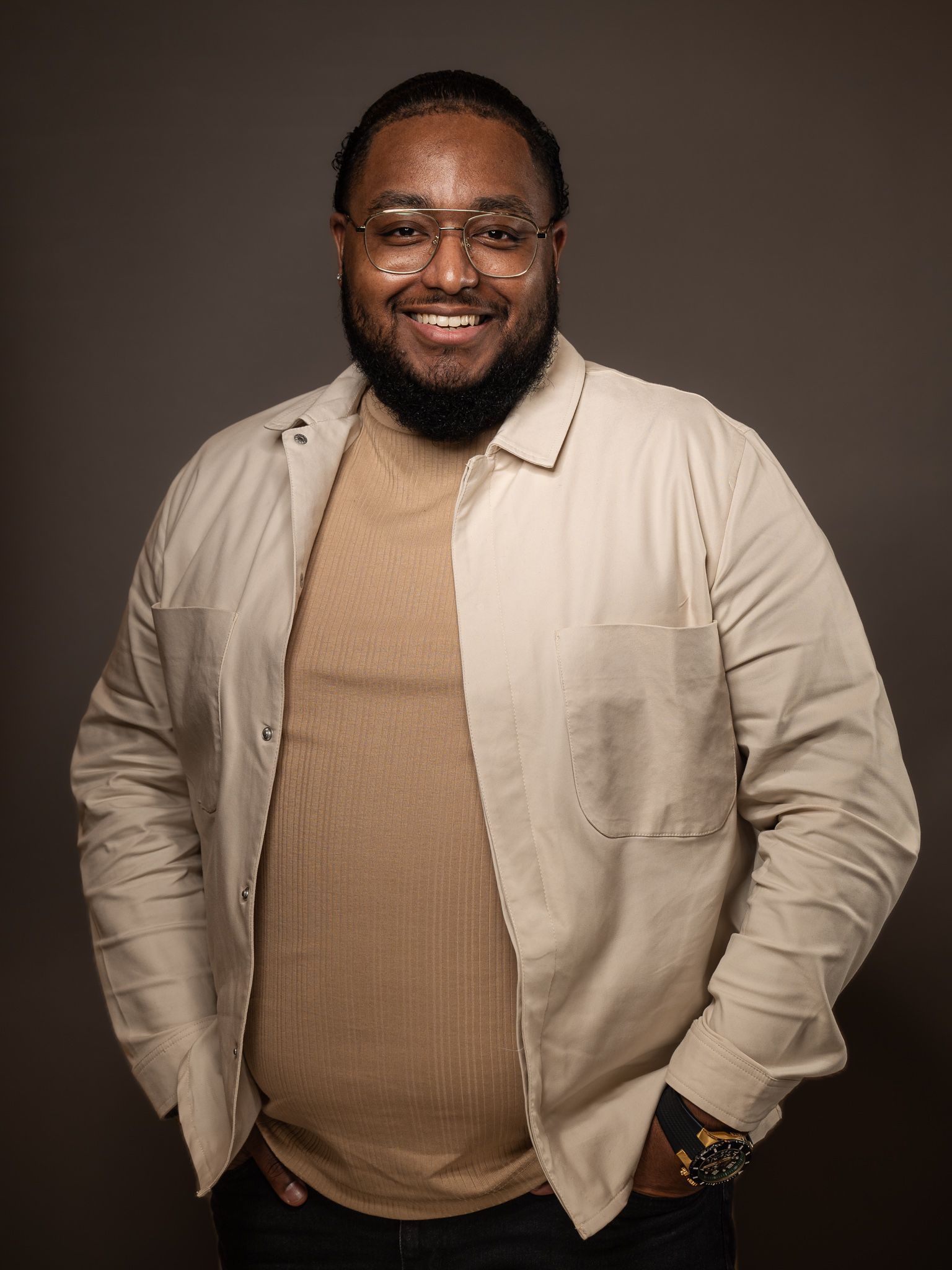 Een man met een baard en een bril staat met zijn handen in zijn zakken en lacht.