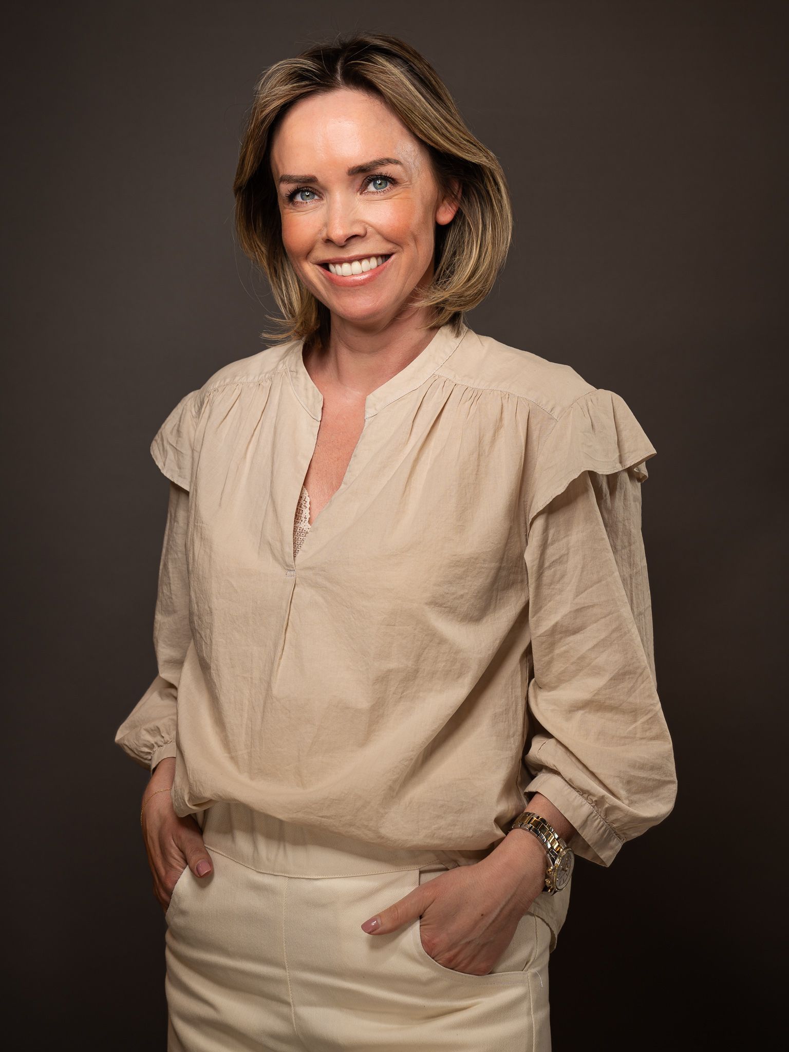 Een vrouw in een beige blouse en een witte broek lacht naar de camera.