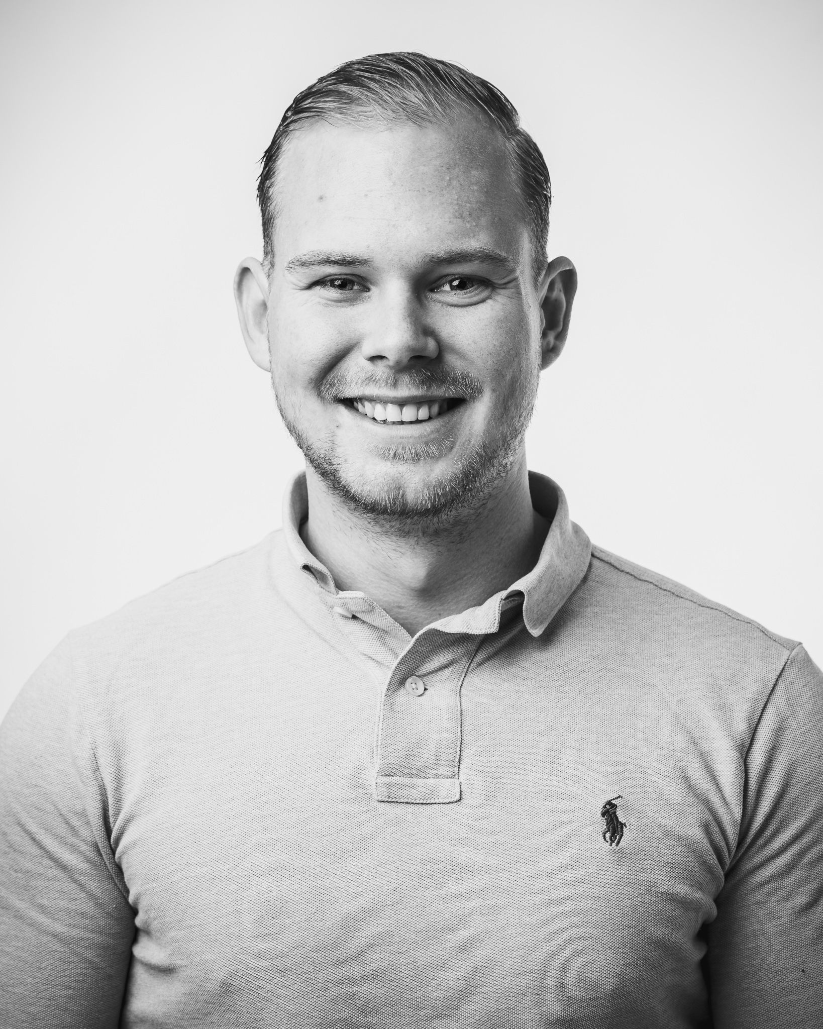Een zwart-witfoto van een lachende man in een poloshirt.