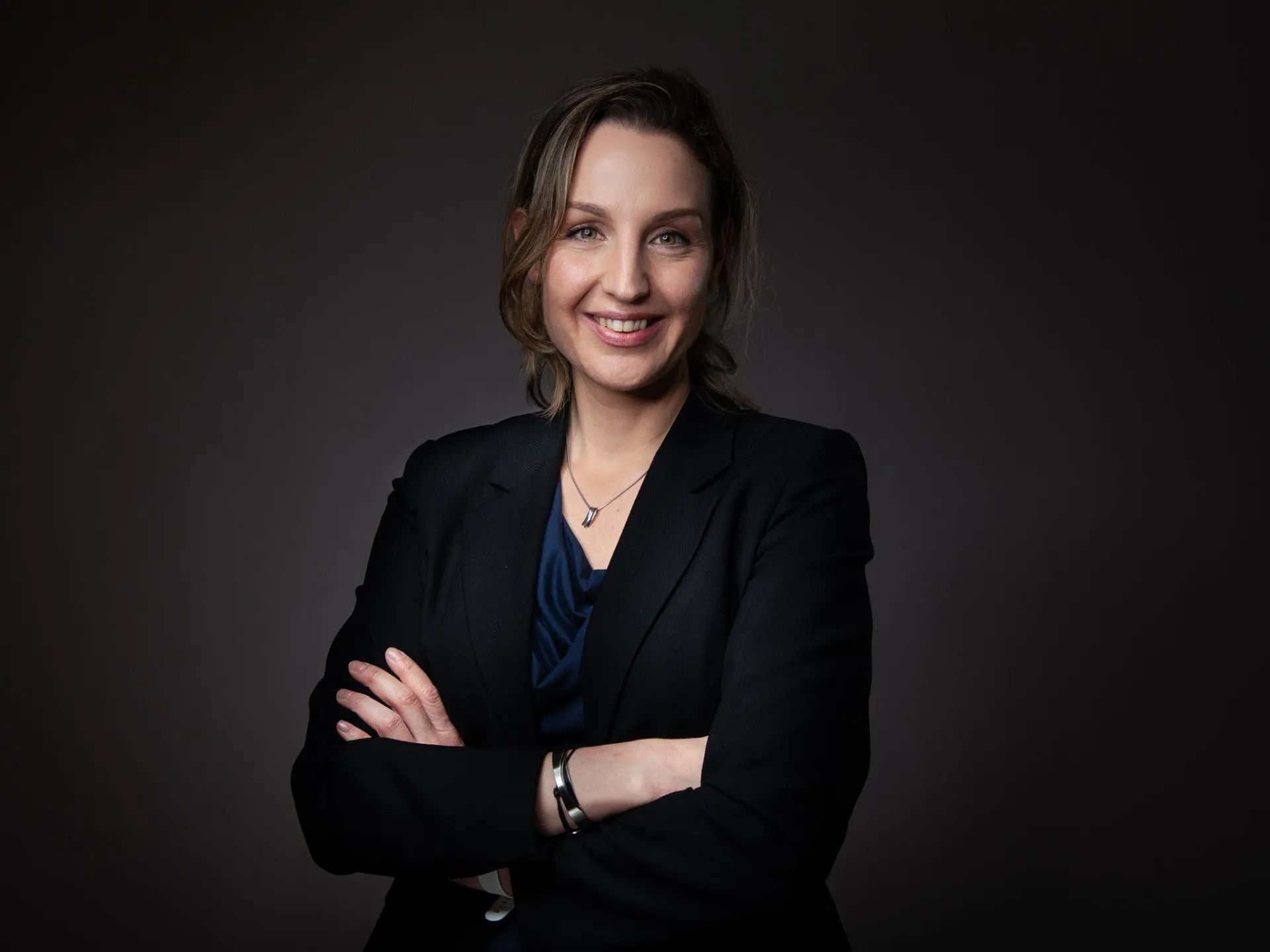 Een vrouw in een pak lacht, met haar armen over elkaar.