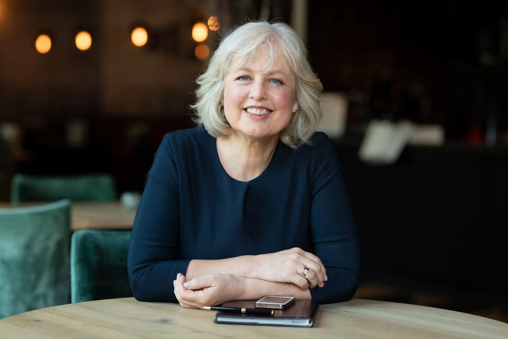 Een vrouw zit aan een tafel, haar armen over elkaar en lacht.