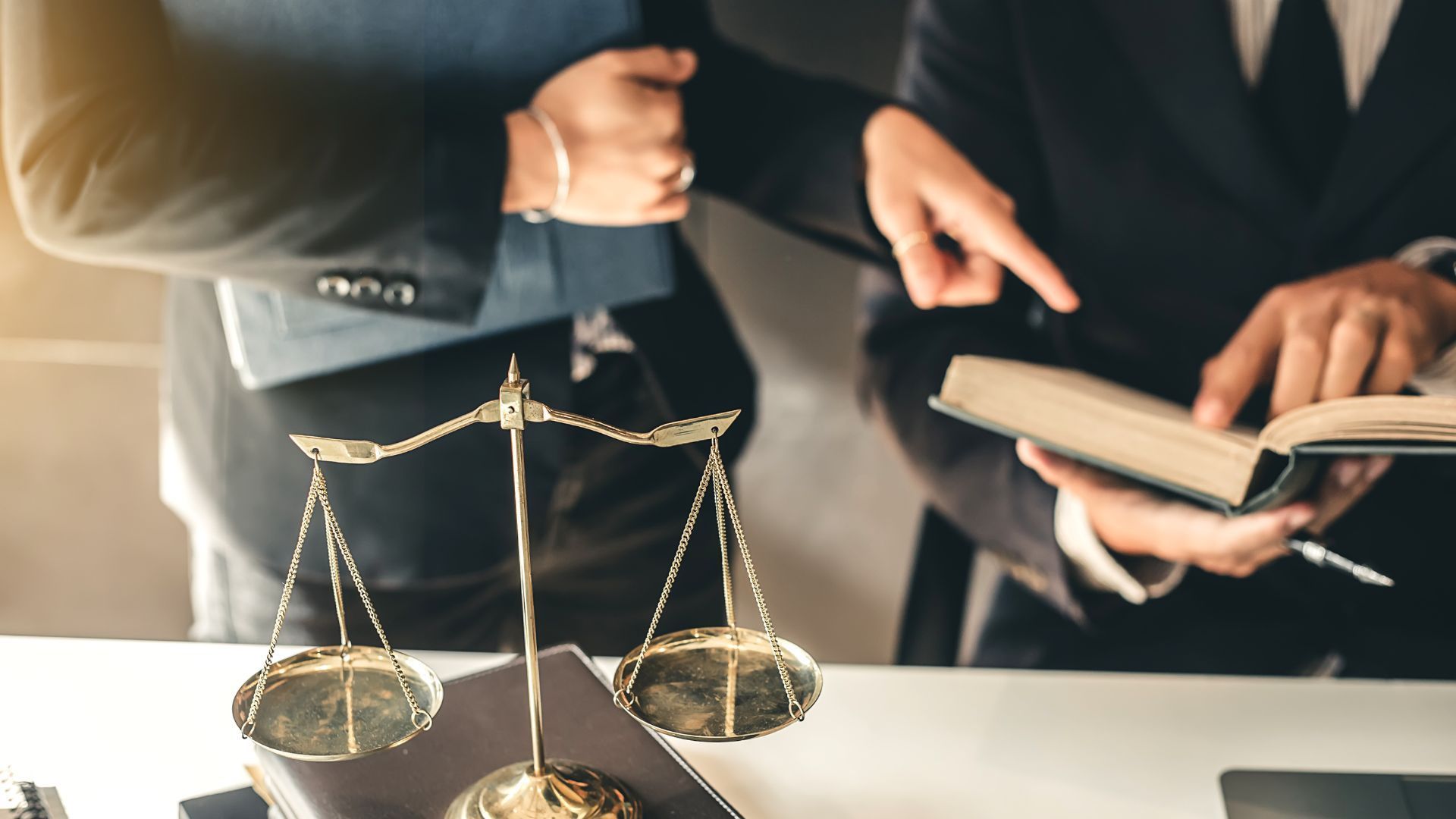 A scale of justice on a desk with two lawyers reviewing a law book.