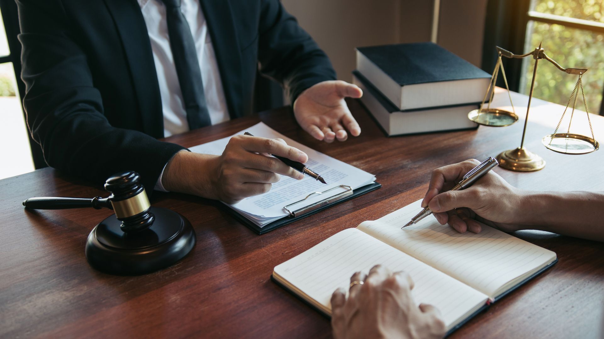 Lawyer explaining documents to a client, both writing at a wooden table, with gavel and scales of justice.