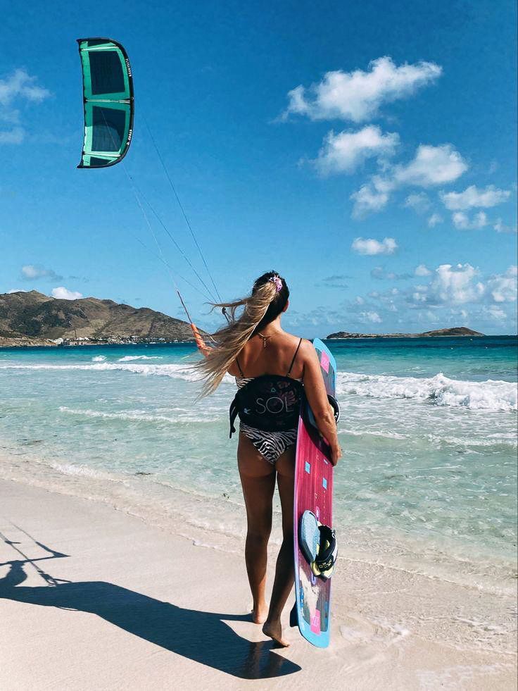 A woman is walking on the beach holding a surfboard and a kite.