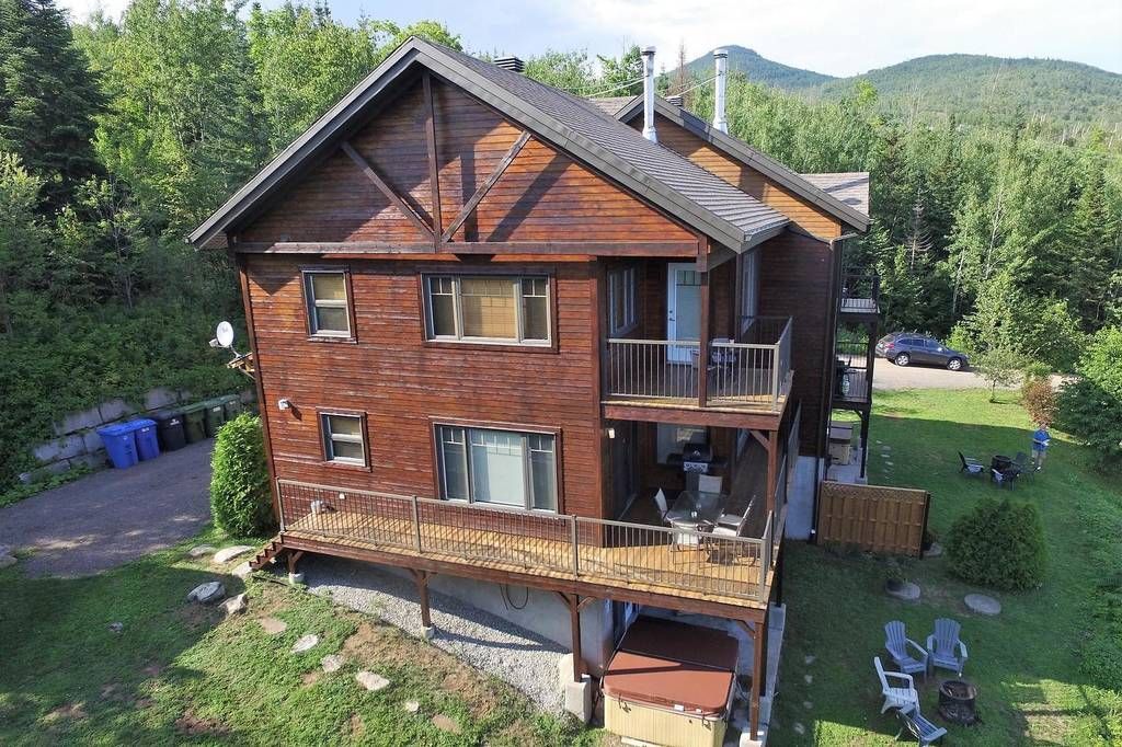 Wooden two-story house with decks and balcony, nestled in a green, wooded area.