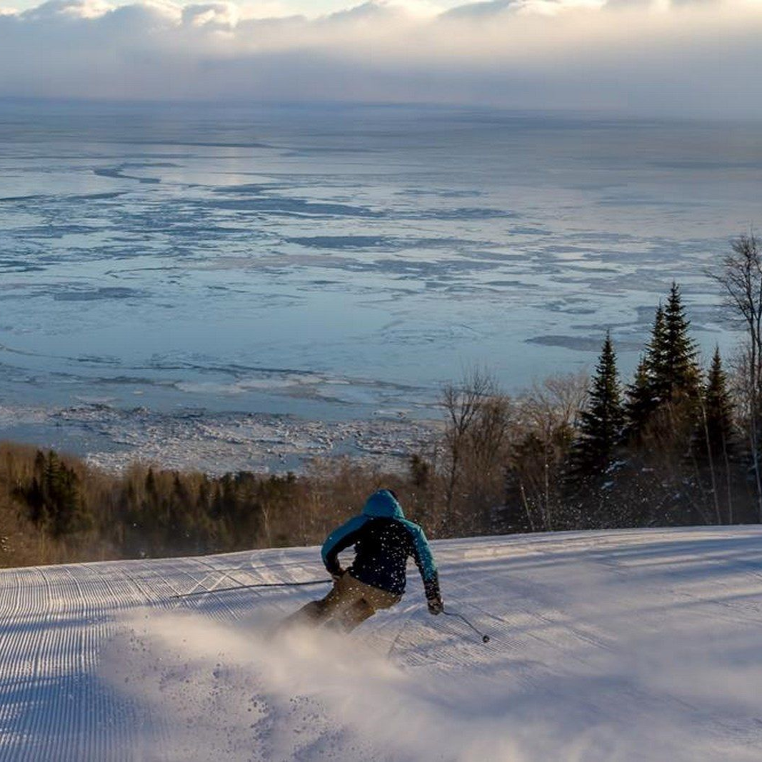 Un skieur dévale une pente enneigée avec un lac gelé en arrière-plan, sous un ciel nuageux.