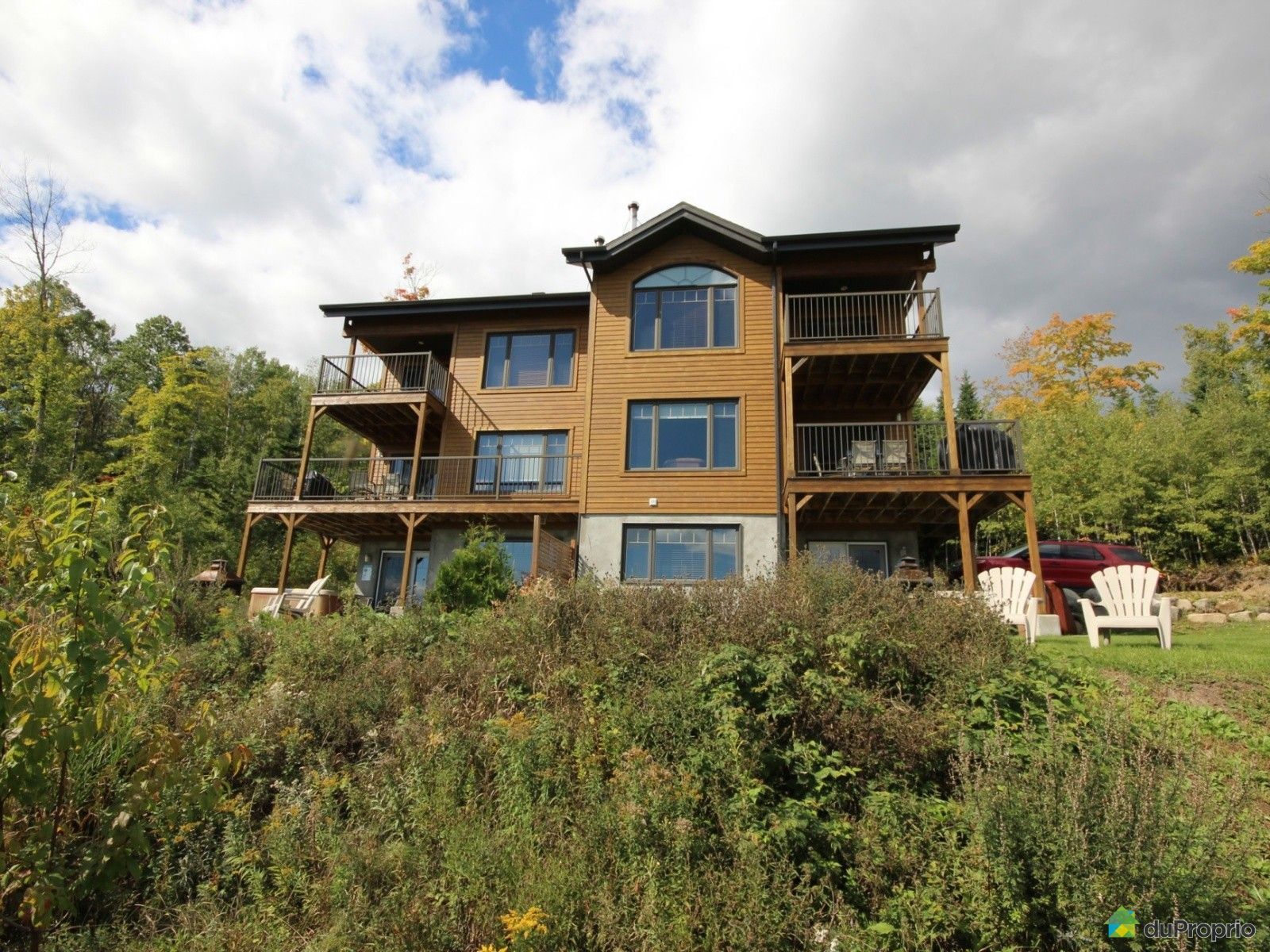 Maison en bois de trois étages avec terrasses donnant sur la végétation et un ciel nuageux.