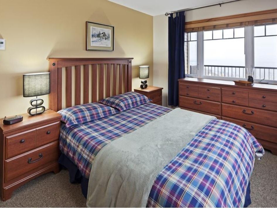 Bedroom with a wooden bed frame, plaid bedding, bedside tables with lamps, and a chest of drawers.