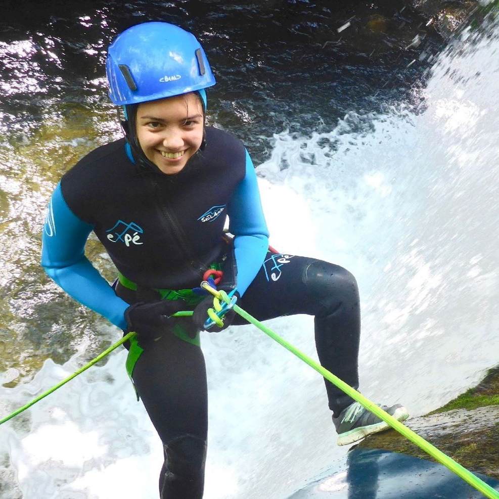 Femme descendant en rappel une cascade, portant un casque bleu et une combinaison de plongée, souriante.