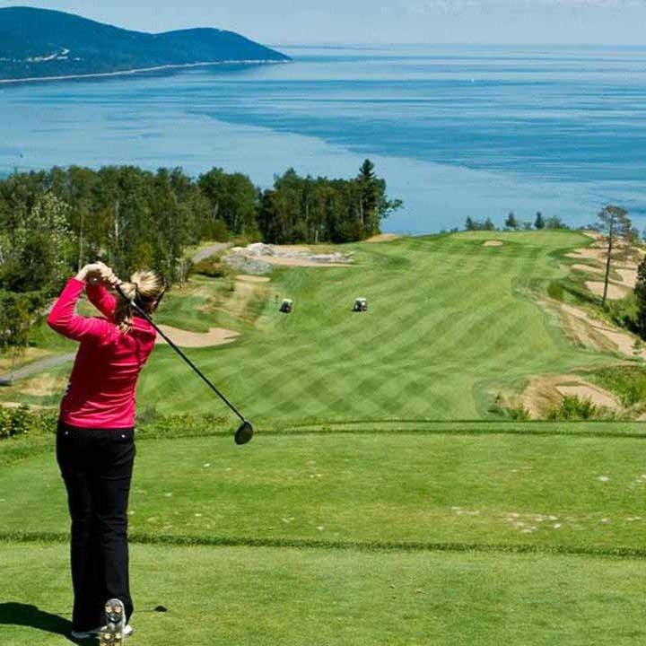 Woman in red swings golf club on a green overlooking ocean and mountains.