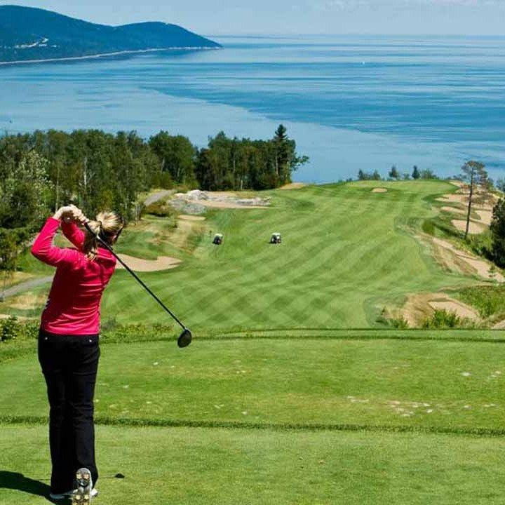 Une femme en chemise rose fait un swing de golf sur un green surplombant une baie pittoresque.