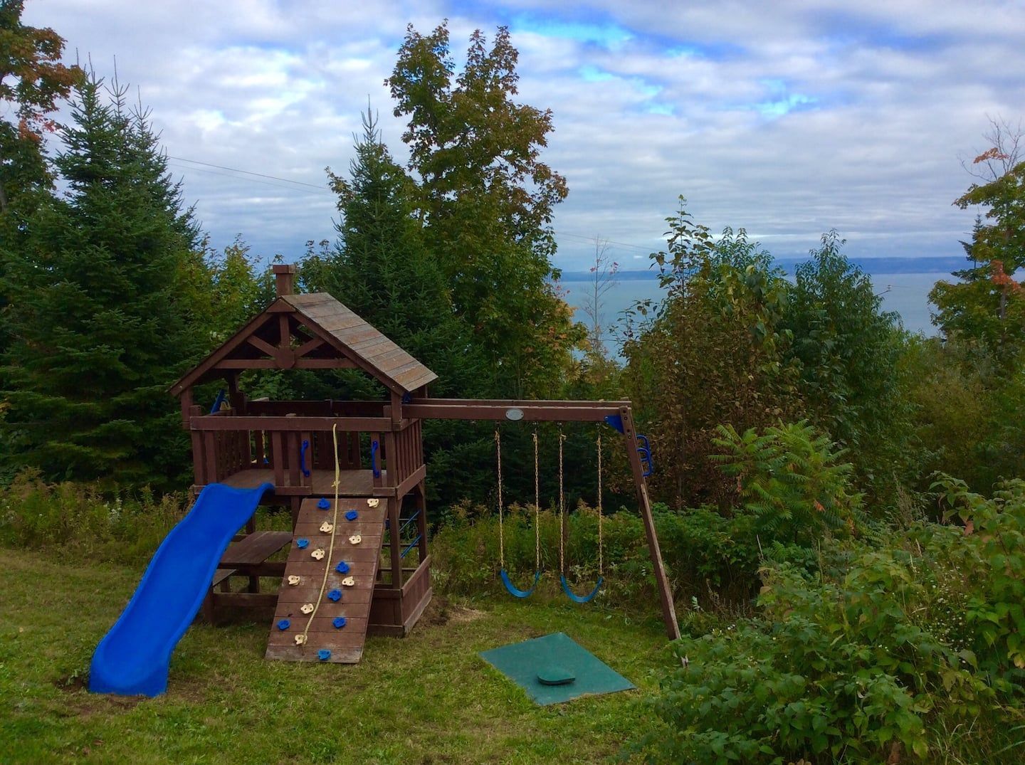 Aire de jeux en bois avec toboggan, mur d'escalade et balançoires, donnant sur un plan d'eau et des arbres.