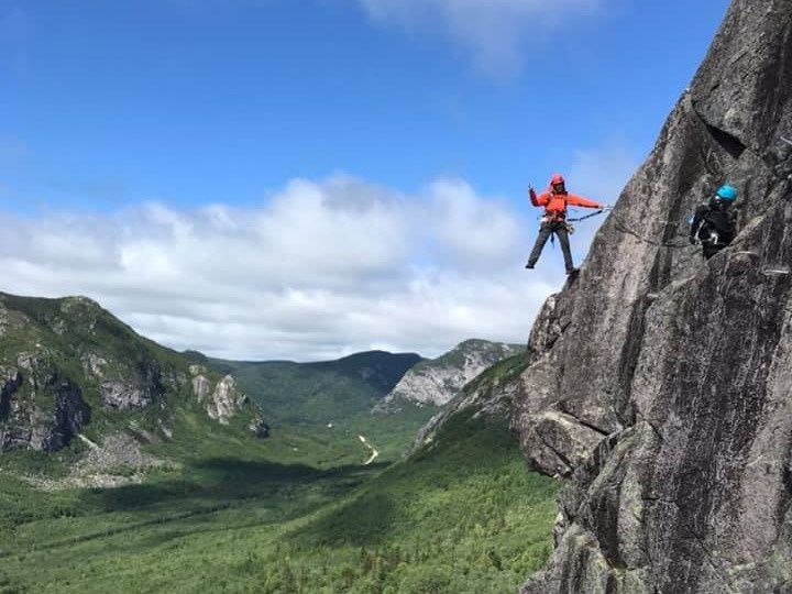 Deux alpinistes sur une falaise rocheuse, l'un d'eux fait signe. Vallée verdoyante en contrebas. Ciel bleu et ensoleillé.