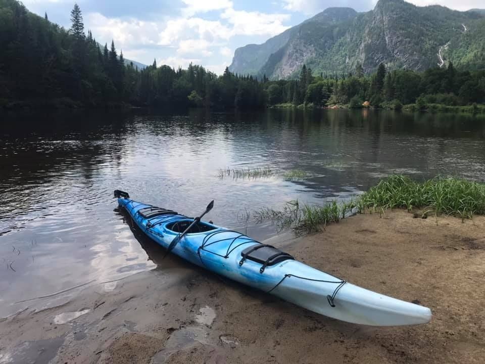 Un kayak bleu sur une rive sablonneuse d'une rivière calme, avec des montagnes et des arbres en arrière-plan.