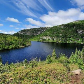 Un lac calme, d'un bleu foncé profond, entouré de collines verdoyantes et boisées, sous un ciel bleu parsemé de nuages.
