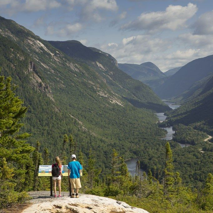 Deux personnes contemplent une vallée pittoresque depuis un promontoire rocheux. La rivière serpente à travers un paysage verdoyant et luxuriant sous un ciel partiellement nuageux.