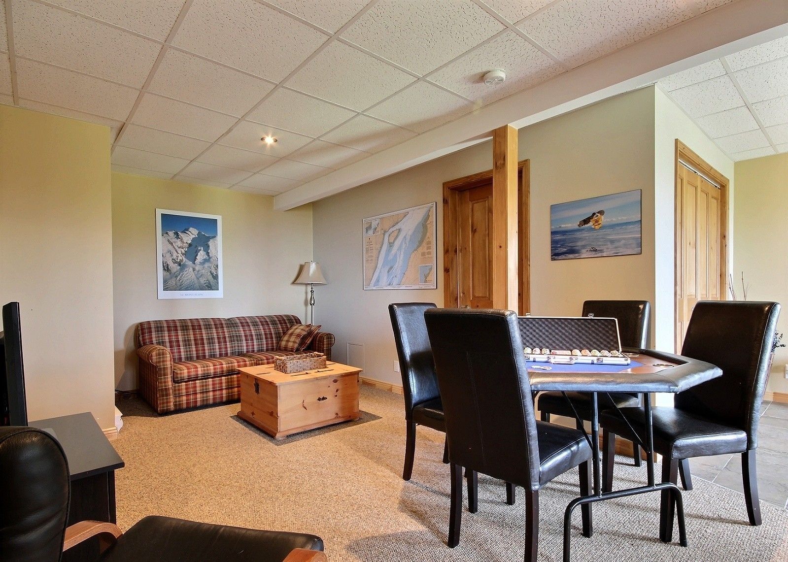 Espace de vie au sous-sol : canapé, table de jeux, œuvres d’art sur des murs beiges, moquette texturée et plafond blanc.