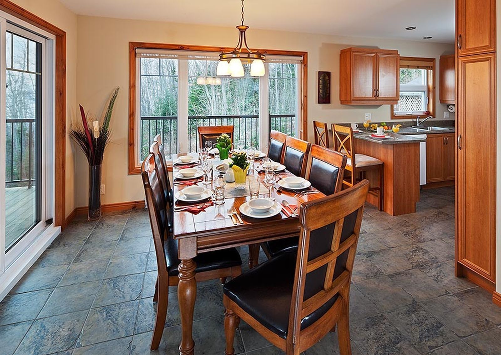Salle à manger avec une longue table en bois dressée pour un repas ; cuisine en arrière-plan, vue sur une terrasse.