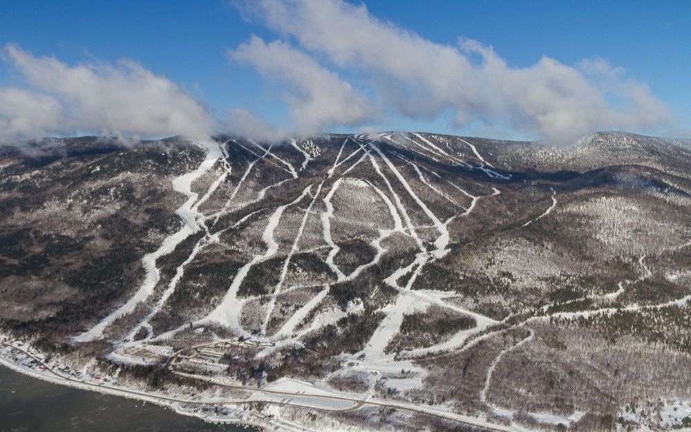 Montagne enneigée avec des pistes de ski tracées à travers les arbres. Ciel bleu parsemé de nuages blancs.