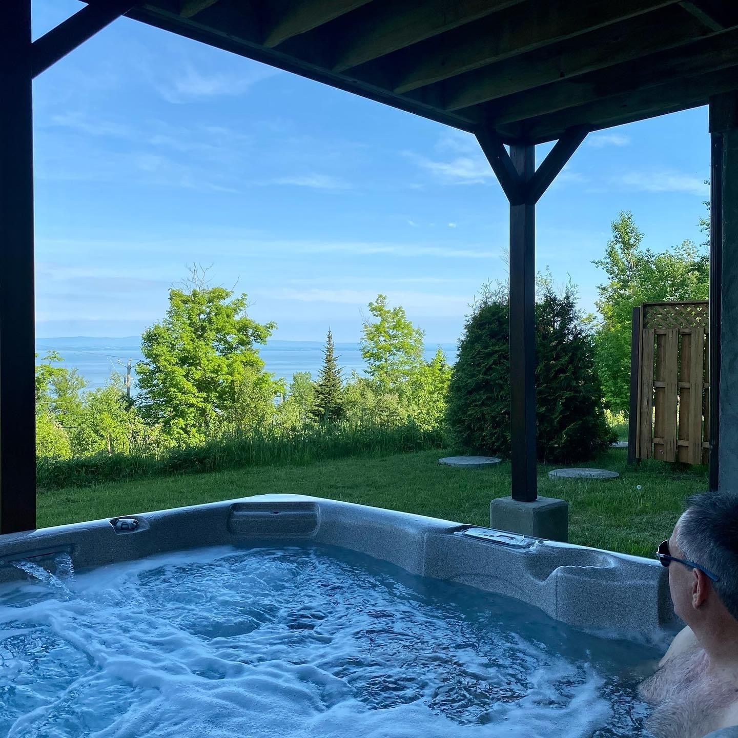 Man in hot tub enjoying scenic view of water and trees under a covered patio.