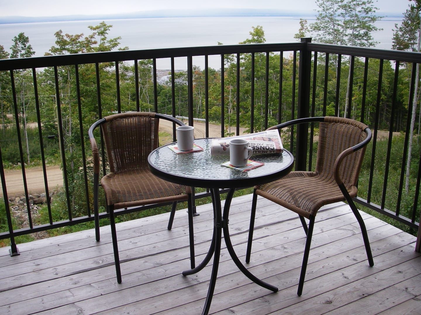 Balcony with two chairs, small table, drinks, and newspaper overlooking a lake.