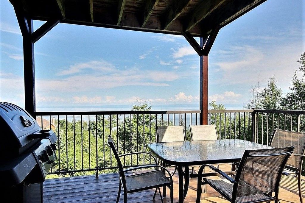 Terrasse avec table, chaises et barbecue donnant sur un plan d'eau et une lisière de forêt sous un ciel d'un bleu éclatant.